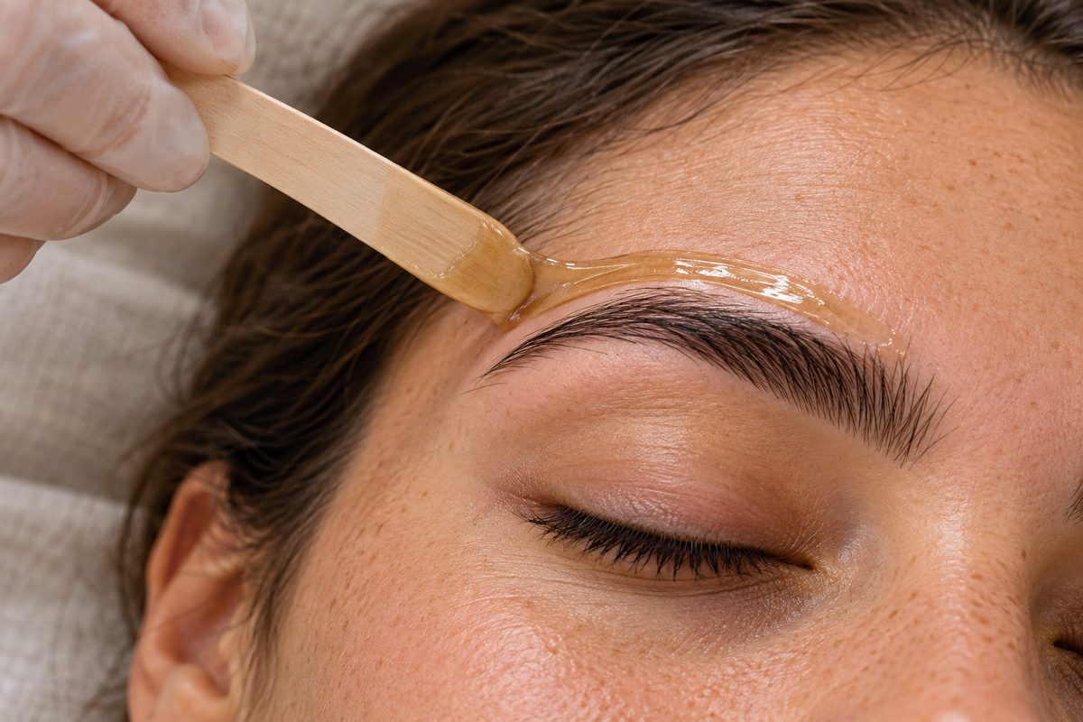 Detailed view of precision eyebrow wax shaping using a wooden applicator and gentle warm wax.